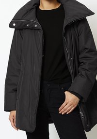 Giacca nera isolante con un ampio colletto, cerniera e chiusure a scatto, e tasche laterali. Indossata sopra un maglione e pantaloni neri.