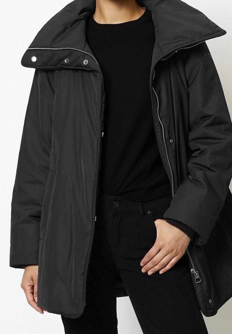 Giacca nera isolante con un ampio colletto, cerniera e chiusure a scatto, e tasche laterali. Indossata sopra un maglione e pantaloni neri.