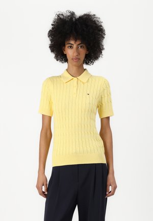 Suéter de polo amarillo de punto con un patrón texturizado, mangas cortas y dobladillo acanalado. Presenta un pequeño logo en el pecho izquierdo.