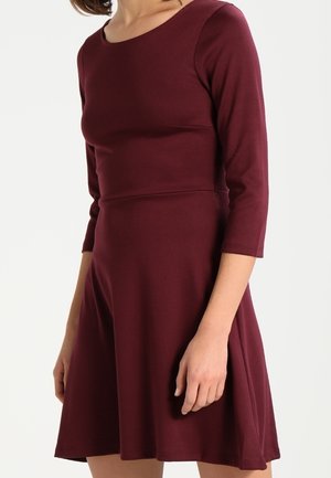 Kobieta w dopasowanej bordowej sukience z rękawami o długości do łokcia i rozkloszowaną spódnicą, stojąca na tle jednolitego białego tła.