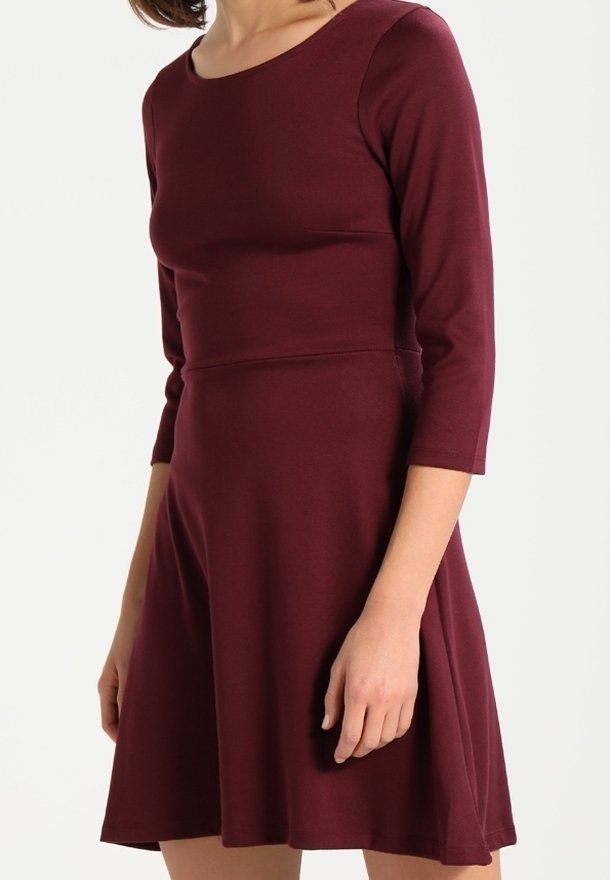 Femme portant une robe bordeaux ajustée aux manches trois-quarts et à la jupe évasée, se tenant devant un fond blanc uni.