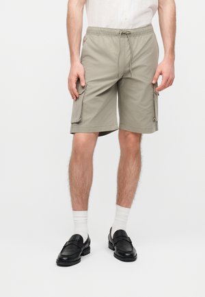 Mand iført beige cargoshorts med snøre i taljen, hvide sokker, sorte loafers og en hvid skjorte med knapper mod en ensfarvet baggrund.