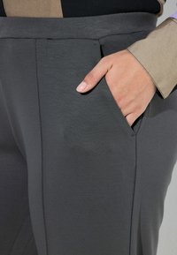 Szare, szyte na miarę spodnie z boczną kieszenią, gładką teksturą i subtelnym połyskiem, prezentujące precyzyjne przeszycia i nowoczesny krój.