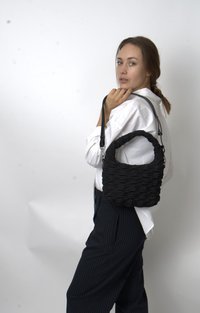 Frau in weißem Hemd und gestreiften Hosen hält eine schwarze geraffte Handtasche über der Schulter und schaut in die Kamera vor einem schlichten Hintergrund.