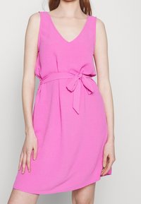 Robe rose sans manches avec un col en V, dotée d'une ceinture nouée, d'une texture douce et d'une coupe évasée, tombant au-dessus du genou.