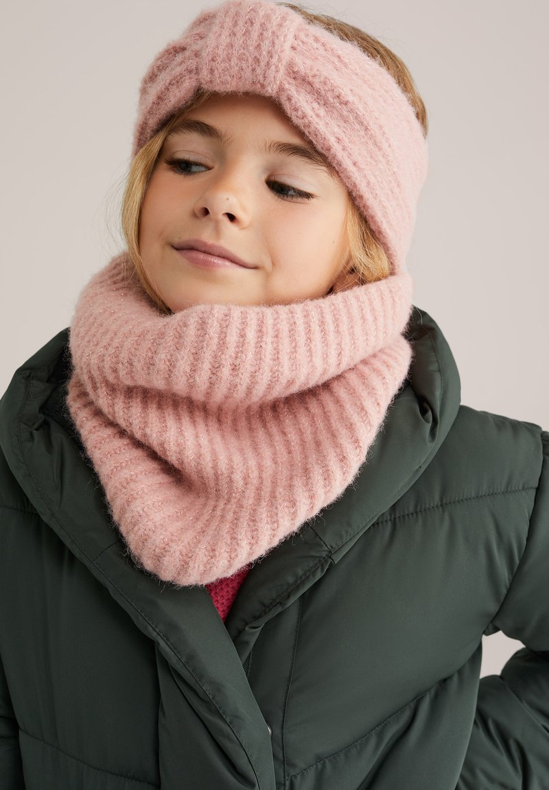 Egy fiatal lány rózsaszín kötött fejpántot és hozzá illő sálat visel, egy sötétzöld pufferek jacket alatt, lágyan mosolyogva, a fejét enyhén megdöntve.