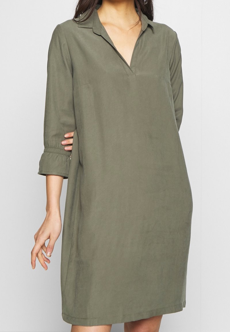 Femme portant une robe longueur genoux vert olive avec col en V et manches trois-quarts, debout devant un fond clair uni.