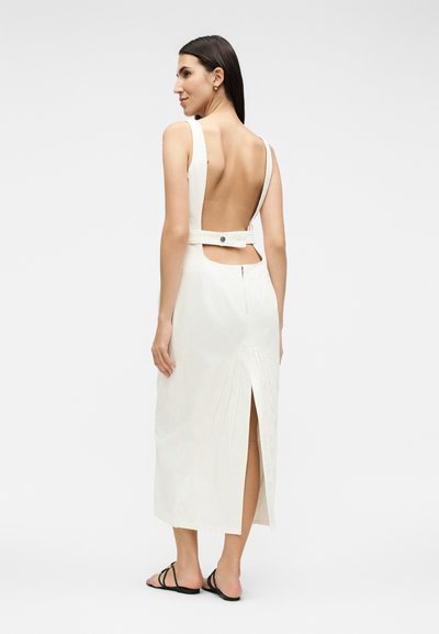 Γυναίκα που φοράει λευκό midi φόρεμα χωρίς μανίκια με ανοιχτή πλάτη, κουμπωτό λουράκι, σκίσιμο στην πλάτη και μαύρα επίπεδα σανδάλια σε απλό φόντο.