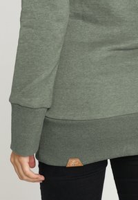 Sudadera verde con puños y dobladillo acanalados, que presenta una pequeña etiqueta de piel marrón en el borde inferior, fabricada con una tela suave.