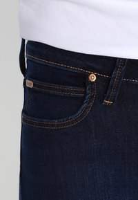 Ciemnoniebieskie jeansy z denimu z mosiężnymi elementami, widocznym przeszyciem i małą metką marki na przedniej kieszeni. Gładka faktura.