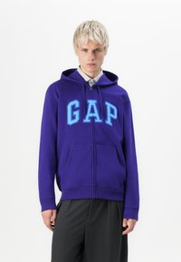 Jonge man met kort blond haar, gekleed in een paarse GAP-rits hoodie over een gestreept shirt en donkere broek, staand tegen een witte achtergrond.