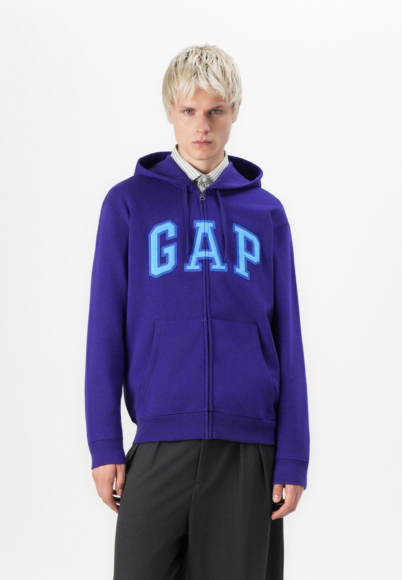 Jonge man met kort blond haar, gekleed in een paarse GAP-rits hoodie over een gestreept shirt en donkere broek, staand tegen een witte achtergrond.