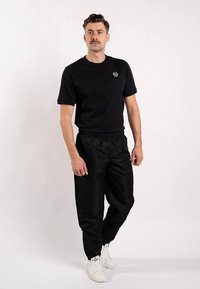 Schwarzes Baumwoll-T-Shirt mit Rundhalsausschnitt und Logo, kombiniert mit schwarzen Hose mit Kordelzug, stehend vor einfachem Hintergrund. Weiße Turnschuhe vervollständigen den Look.