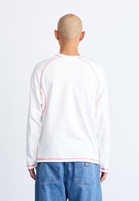 White long-sleeve shirt with red stitching accents and a rounded neckline, paired with blue denim pants.