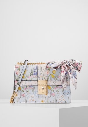 Handtas met bloemenpatroon, gouden sluiting, kettingriem en bijpassend stoffen strikje, geplaatst op een wit voetstuk.