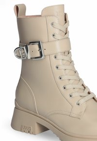 Stivaletto in pelle beige con dettaglio in fibbia argentata, chiusura con lacci e tacco piatto. Presenta una texture liscia e sottili accenti di cucitura.