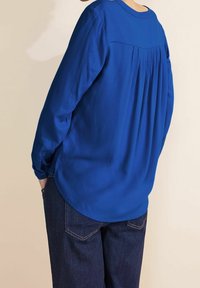 Blusa blu a maniche lunghe con un design sul retro a pieghe e scollatura rotonda, abbinata a jeans scuri. Tessuto liscio, vestibilità casual.