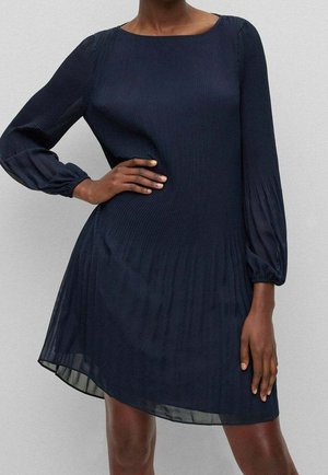 Vrouw draagt een marineblauwe geplooide jurk met lange doorschijnende bischofsmouwen, één hand op de heup, de andere langs haar zijde.