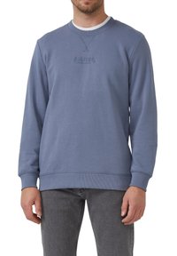 Muž v modrom svetríku s okrúhlym výstrihom so subtilným embosovaným logom "S. Oliver" a tmavohnedých džínsoch, stojí proti bielej stene.