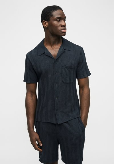 Homme portant une chemise à manches courtes boutonnée bleu marine foncé et un short assorti avec une texture en chevrons, regardant vers sa gauche sur un fond uni.