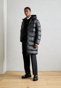 Zwarte puffercoat met een capuchon, met een glanzende buitenkant, ritssluitingen en een horizontaal gewatteerd patroon. Gedragen over een zwarte trui en broek.