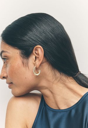 Woman with dark hair tied back, wearing gold hoop earrings and a dark sleeveless top, looking to the left against a plain background.