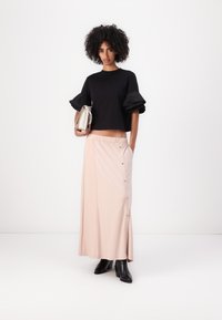 Schwarzes Crop-Top mit Puffärmeln, kombiniert mit einem hellrosa Maxirock mit seitlichen Knöpfen und Taschen, getragen mit schwarzen Stiefeletten mit Absatz.