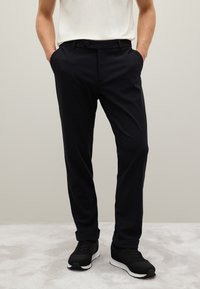 Pantaloni neri slim fit, con tasche laterali e chiusura a un bottone. Indossati con sneakers nere su una superficie grigia chiara.