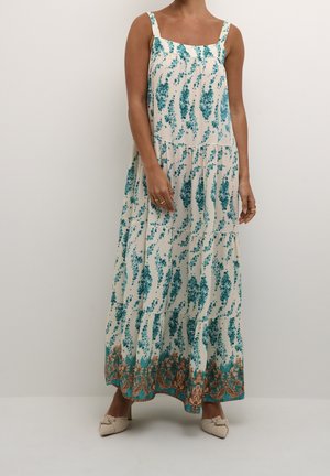 Kvinde iført hvid ærmeløs maxikjole med blå blomsterprint og ornamenteret blå og gylden kant nær kanten, kombineret med beige slip-on sko.