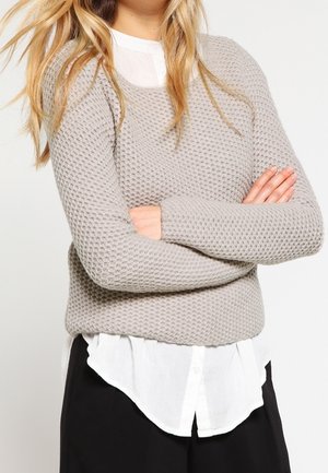 Frau mit langen blonden Haaren, trägt einen grauen strukturierten Pullover über einem weißen Hemd mit Knöpfen, die Arme verschränkt, gekleidet in schwarze Hosen.