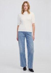 Witte, fuzzy, kort mouwsweater met een hoge halslijn, gecombineerd met lichtblauwe, wijde jeans. Zwarte, puntige boots maken de outfit compleet.