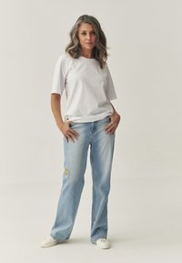 Hvid kortærmet t-shirt parret med lyseblå jeans, der har gule og røde broderede detaljer. Modellen står med hænderne på hoften.