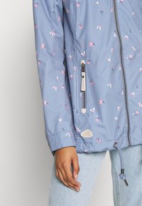 Veste bleue avec imprimé floral rose et blanc, fermeture éclair devant, poche latérale zippée, ourlet à cordon, portée avec un jean bleu clair sur le mannequin.