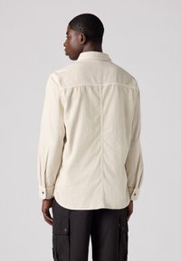 Light beige long-sleeve shirt with a collar and button cuffs, featuring a stitched seam down the back and a relaxed fit.