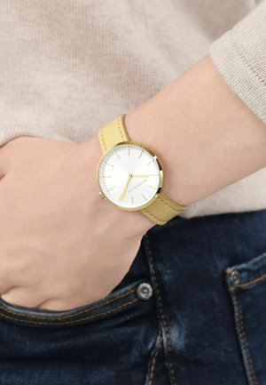 Armbanduhr mit goldenem Gehäuse und gelbem Lederarmband am linken Handgelenk, getragen mit beigem Pullover und dunkelblauer Jeans mit sichtbarem Knopf und Naht.