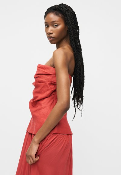 Hosszú, fonott haját viselő nő pánt nélküli, ráncolt piros ruhában, oldalnézetben, egyszerű világos háttér előtt áll.