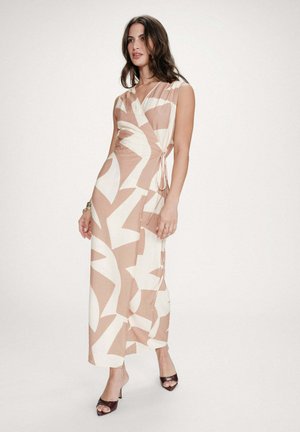 Femme debout portant une robe portefeuille sans manches beige et blanche à motif abstrait avec un lien sur le côté, associée à des chaussures à talons ouvertes foncées sur un fond uni.