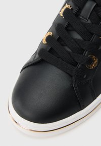 Μύτη μαύρου δερμάτινου αθλητικού παπουτσιού με χοντρά μαύρα κορδόνια περασμένα από χρυσά πλεκτά, λευκή σόλα με λεπτή χρυσή ρίγα.