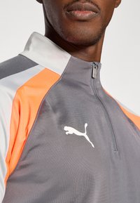 Šedý sportovní sako se zipovým límcem, s oranžovými a bílými akcenty. Tkanina má hladkou texturu, na hrudi je bílé logo Puma.