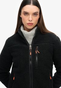 Zwarte fleecetrui met een hoge kraag, een gestructureerd oppervlak en contrasterende ritsen met oranje accenten op de zakken en de hoofdrits.