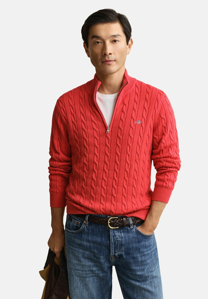 Uomo con maglione rosso a trecce con mezza cerniera indossato sopra una camicia bianca, jeans blu, cintura intrecciata marrone, che tiene in mano una giacca marrone.