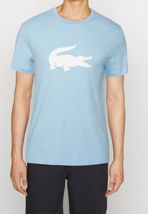 Person trägt ein hellblaues Kurzarm-T-Shirt mit einer großen weißen Krokodil-Grafik auf der Brust und dunkle Hose.