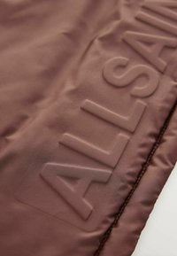 Brun tyg med en slät textur som har en präglad "ALLSAINTS"-logotyp. Designen inkluderar subtila sömmar längs kanterna.