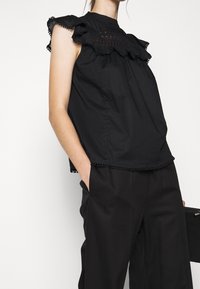 Mujer vestida con una blusa sin mangas negra con panel de encaje con volantes y pantalones negros, con la mano en el bolsillo, sosteniendo un bolso de mano negro.