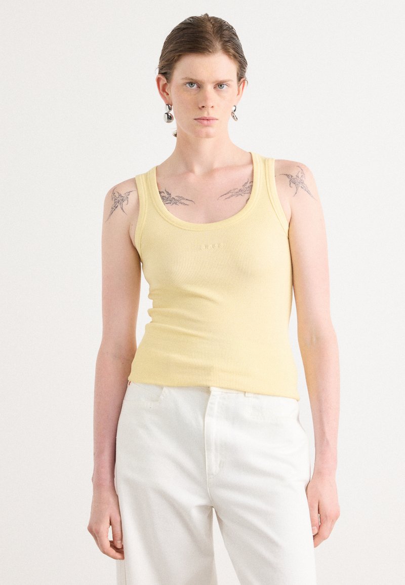 Yellow ribbed tank top with a scoop neck, fitted shape. Worn with white trousers, featuring visible tattoos on the shoulders. Silver earrings present.