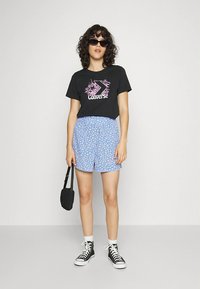 Camiseta preta com logótipo Converse rosa, calções florais azul claro, mala preta e ténis pretos de cano alto com solas e atacadores brancos.