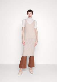 Abito lavorato a maglia beige con un motivo geometrico, sovrapposto a una maglietta bianca a maniche corte, abbinato a pantaloni larghi marroni e scarpe con plateau nude.