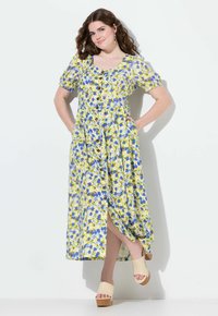 Žluté květinové midi šaty s nafouknutými rukávy, předními knoflíky a bočními kapsami; zahrnují rozparek a jsou doplněny béžovými platformovými sandály.