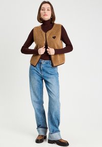 Brun fleece vest med en hjerteformet lap, båret over en mørk rullekrave, parret med lyseblå jeans og sko med leopardmønster.