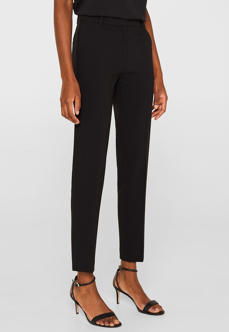 Personne portant un pantalon noir ajusté sur mesure et des sandales à talons hauts ouvertes sur un fond uni.